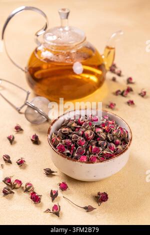 Getrocknete Rosenwürfel füllen eine weiße Schale, bereit zum Aufbrühen neben einem Glas mit Teekanne mit goldenem Tee und einem metallischen Aufgusszubehör, symbolisch für Kräuterwohlbefinden Stockfoto
