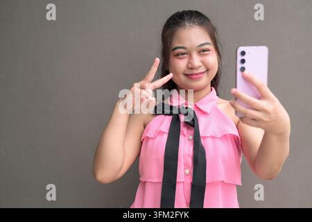 Ein mittleres Porträt einer jungen südostasiatischen thailändischen Frau, die ein rosa Smartphone hält, um ein Selfie zu machen und dabei ein Friedenszeichen zeigt. Lässige, moderne Stimmung aga Stockfoto