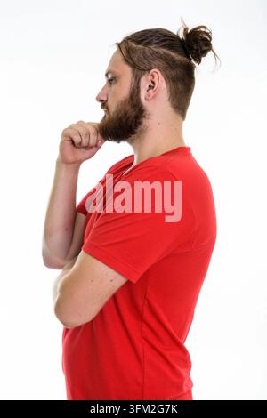 Seitenprofil-Studio-Porträt eines bärtigen jungen kaukasiers mit einem Haarbutt, der ein rotes T-Shirt mit V-Ausschnitt trägt. Er posiert in einer denkenden Geste mit ha Stockfoto