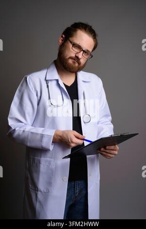 Studio-Porträt eines bärtigen jungen männlichen Arztes in einem weißen Labormantel mit Stethoskop, mit Klemmbrett und Stift. Medizinisches Fachpersonal Rev Stockfoto