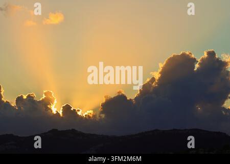 Eine spektakuläre Sonnenuntergang- oder Sonnenaufgangsszene mit intensiven goldenen Krepuskularstrahlen, die hinter dunklen, schweren Kumuluswolken platzen und die Hügelkuppen umrahmen Stockfoto
