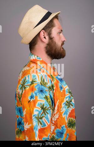 Seitenansicht eines jungen kaukasischen Mannes mit Bart, der ein leuchtend orangefarbenes Hawaiihemd mit Blumenmuster und einen Trilby-Strohhut trägt. Ideal für Reisen, Urlaub und Stockfoto