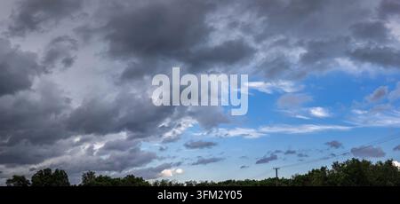 Gewitterwolken, die einen Teil des Himmels bedecken, bilden einen starken Kontrast zum klaren blauen Himmel und der Reihe grüner Bäume am Horizont, was den Wandel darstellt Stockfoto