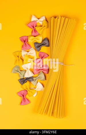 Trockene Farfalle-Pasta in verschiedenen Farben neben einem gebundenen Bündel Spaghetti auf gelbem Hintergrund, lebhaftes Stillleben von ungekochtem itali von oben Stockfoto
