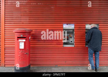 Ein Mann hebt Geld an einem Geldautomaten vor dem geschlossenen Hanwell Town Centre Post Office im Westen Londons ab, während die Filiale geschlossen ist. Stockfoto