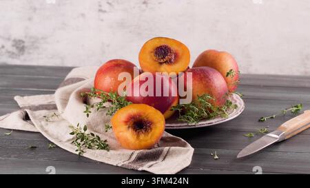Nahaufnahme von reifen frischen Pfirsichen, garniert mit aromatischen Thymianzweigen auf einem rustikalen Teller und Stofftuch neben einem kleinen Messer auf einem dunklen Holztisch Stockfoto