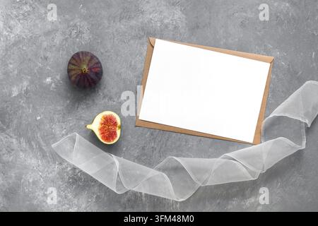 Feminines Hochzeitseinladungsdesign. Leere weiße Karte als Mocap, Feige, Band, Bastumschlag. Grauer Betonhintergrund. Minimalistische Schreibwaren-Szene. Oberansicht, Flat Lay Stockfoto