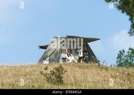 Familie entspannt in Campingstühlen vor ihrem Zelt auf einem grasbewachsenen Hügel unter einem klaren blauen Himmel, Ta Xua, Son La, Vietnam Stockfoto