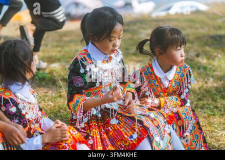Drei Mädchen in bestickten Hmong-Kostümen sitzen auf dem Gras, während eines mit silbernen Münzornamenten spielt, Ta Xua, Son La, Vietnam Stockfoto