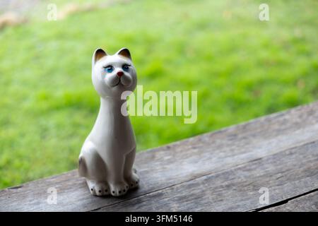 Eine keramische Katzenfigur mit blauen Augen sitzt auf einer verwitterten Holzbank neben einem grünen, grasbewachsenen Garten und schafft eine ruhige Außenszene mit neugierigem Ausdruck a Stockfoto