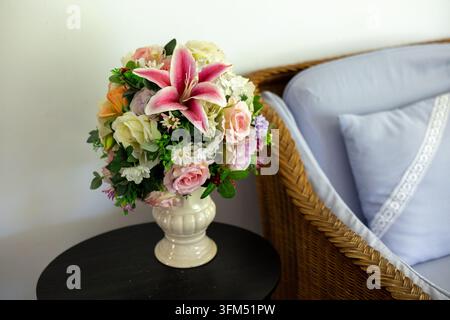 Ein Blumenarrangement mit rosa Lilie und Rosen in einer Keramikvase neben einem Korbstuhl, das eine sanfte Innendekorationsszene mit frischen Farben und Texturen schafft Stockfoto