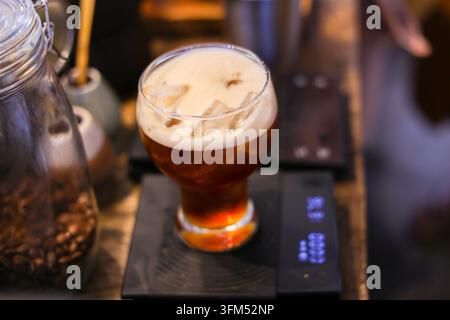 Ein Eiskaffee im Glas steht auf einer digitalen Waage neben Kaffeebohnen und schafft eine warme Cafészene mit cremigem Schaum und reicher brauner Farbe Stockfoto