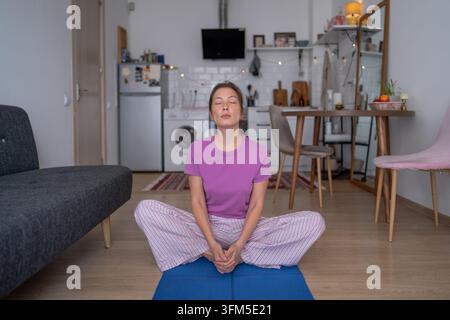 Bewusste Pause einer Frau, die zu Hause Meditation praktiziert, sitzt auf einer Matte mit geschlossenen Augen zur Stressbewältigung Stockfoto