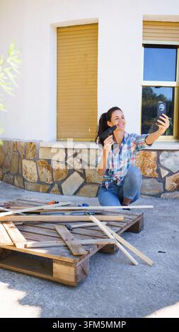 Frau filmt sich selbst mit einem Smartphone, während sie an einem Holzpaletten-Projekt arbeitet Stockfoto