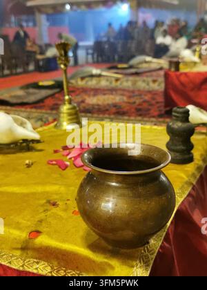 VARANASI, INDIEN - 23. JANUAR 2026: Ein Messingritualtopf und Rosenblätter ruhen auf einem gelben Tuch vor der Morgendämmerung Aarti am Assi Ghat. Stockfoto