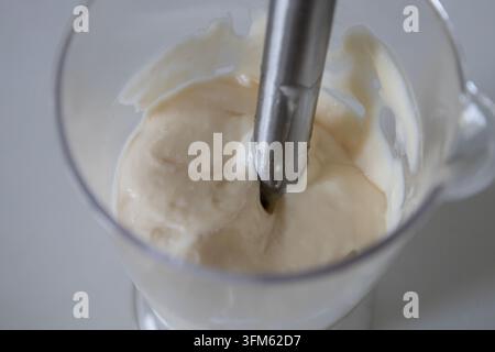 Hausgemachte Mayonnaise oder Sauce mit einem Handmixer in einem durchsichtigen Plastikbecher zubereiten, cremige weiße Zutaten für ein Rezept in der k Stockfoto