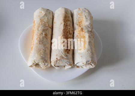 Drei frisch zubereitete hausgemachte Shawarma-Wraps mit goldener, gerösteter Lavash-Kruste, die ordentlich auf einem weißen Keramikteller gegen einen sauberen Hintergrund liegen, oben drauf Stockfoto