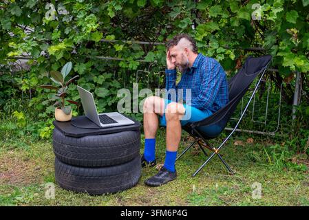 Bärtiger Mann in einem Klappstuhl arbeitet an einem Laptop, der auf gestapelten Reifen in einem grünen Hinterhof platziert ist, und wirkt gestresst und konzentriert. Stockfoto