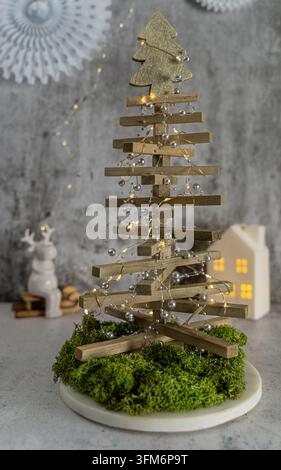 Moderner minimalistischer Weihnachtsbaum aus Holzlatten mit Lichterkette auf einem moosbedeckten Sockel und einem leuchtenden dekorativen Haus im Hintergrund Stockfoto