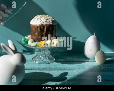Traditionelles Paska Pasca-Osterbrot, serviert auf einem gläsernen Kuchenständer mit dekorativen Ostereiern und einem weißen Hasenornament auf einem türkisfarbenen Holz Hintergrund Stockfoto