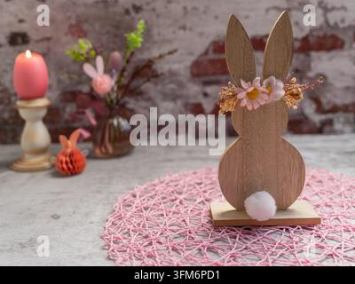 Holzener Osterhase als Ornament, dekoriert mit Frühlingsblumen und einem weißen dekorativen Schwanz auf einer rosa texturierten Matte vor einem rustikalen Ziegelhintergrund Stockfoto
