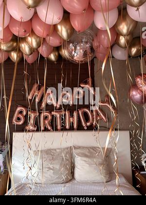 Metallische Geburtstagsbuchstaben und Luftballons schmücken ein helles Schlafzimmer im warmen Sonnenlicht. Stockfoto