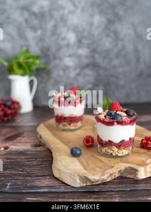 Zwei individuelle geschichtete Mascarpone-Desserts mit frischen Beeren und knuspriger Granola, serviert in Gläsern auf einem rustikalen Holzbrett Stockfoto