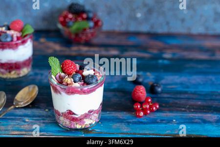 Individuelles geschichtetes Mascarpone-Dessert mit frischen Beeren und knuspriger Granola, serviert in einem Gläser auf einem rustikalen blauen Holzuntergrund Stockfoto