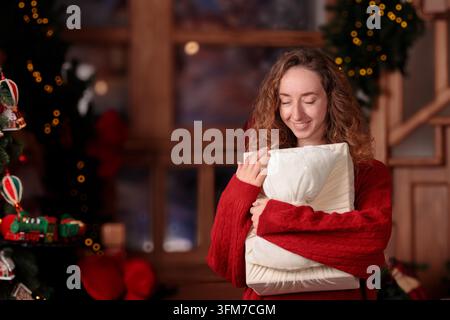 junge Frau mit lockigem Haar trägt einen warmen roten Pullover, während sie ein Geschenk in der Nähe eines leuchtenden Baumes während einer friedlichen festlichen Feier umarmt Stockfoto
