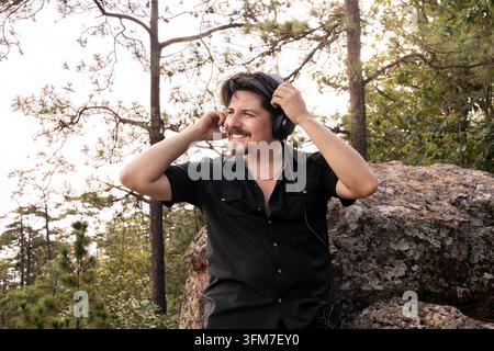 Ein gutaussehender, lächelnder junger Mann in einem schwarzen Hemd stellt seine Kopfhörer ein, während er an einem Felsen in einem Kiefernwald lehnt. Stockfoto