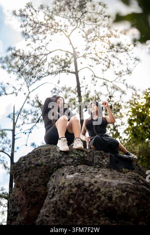 Eine Niederwinkelaufnahme von zwei jungen Freundinnen, die auf einem großen Felsen in einem Bergwald sitzen und Musik über Over-Ear-Kopfhörer hören; sie haben Stockfoto
