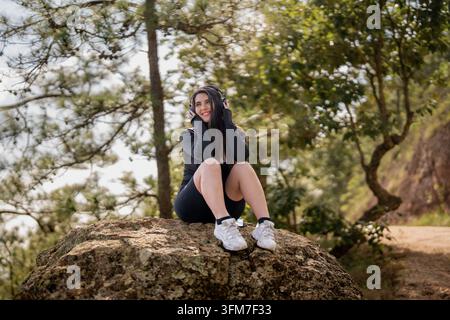 Eine attraktive junge Frau in schwarzer Sportkleidung, lächelnd und ihre Over-Ear-Kopfhörer haltend, sitzt auf einem großen Felsen im Wald. Stockfoto