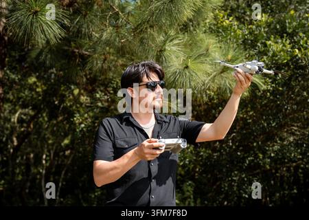 Ein gutaussehender junger Mann mit langen dunklen Haaren, der eine Sonnenbrille und ein schwarzes Hemd trägt, hält eine Drohne zum Abflug mit der Fernbedienung im Wald. Stockfoto
