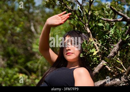 Nahaufnahme-Porträt einer jungen Frau in Schwarz, die an einem Baum lehnt und ihr Gesicht mit der Hand vor der Sonne schützt im Wald Stockfoto