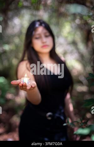 Nahaufnahme einer Hand, die eine kleine brennende Kerze hält, mit unscharfer junger Frau im Wald Stockfoto
