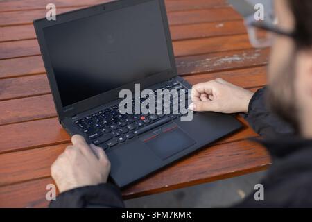 Eine teilweise sichtbare Person verwendet einen offenen Laptop auf einem hölzernen Außentisch. Die nahe Ansicht von oben zeigt Hände, Tastatur, leeren Bildschirm und flache Hintergrundbeleuchtung Stockfoto