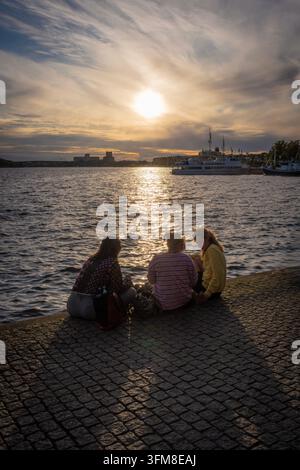 Drei junge Frauen sitzen auf einem Steinpier und genießen einen Snack am Wasser bei Sonnenuntergang mit Booten und einem leuchtenden Himmel, der sich im Hafen von Stockholm spiegelt Stockfoto