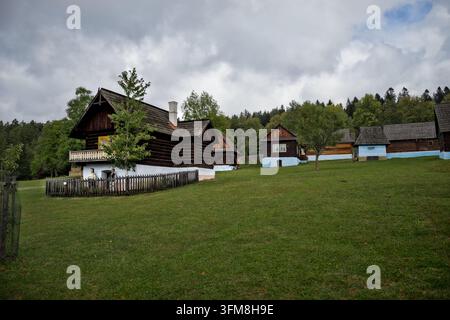 Historische Holzhäuser mit weiß-blau gestrichenen Sockeln stehen auf einem grasbewachsenen Hügel nahe einem Wald unter einem bewölkten Himmel. Stockfoto