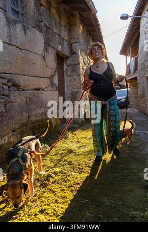 Zufriedene schwangere Frau genießt einen Spaziergang mit ihren beiden Hunden an einem sonnigen Nachmittag in einer traditionellen Stein-Dorfstraße Stockfoto