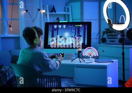 Spielsucht und digitale Unterhaltung, Teenager spielt Videospiele am Computer mit Controller und Kopfhörern in einem neonbeleuchteten Raum in der Nacht Stockfoto