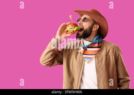 Fast-Food-Vergnügen und Junk-Food-Heißhunger, Mann mit Cowboyhut isst mit Appetit einen Burger vor einem leuchtend rosa Hintergrund Stockfoto