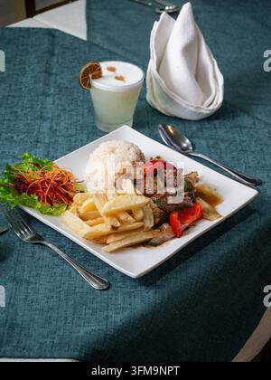 Weißer Teller enthält Reis, Pommes, Steak und Gemüse. Ein Teeglas mit Zitrone steht neben einer ordentlich gefalteten Serviette. Besteck liegt neben dem Gericht auf dem blaugrünen Tisch Stockfoto