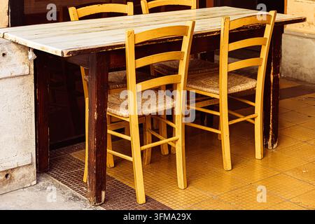 Ein Außenrestaurant-Setup mit einem rustikalen Holztisch, gepaart mit mehreren klassischen gelben Stühlen mit Strohsitzen. Stockfoto