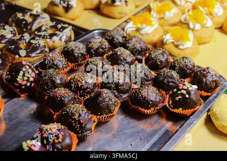Schokoladenkugeln mit bunten Streuseln in orangefarbenen Pappbechern auf einem Metalltablett, dahinter verschiedene Donuts mit Toppings in Thailand. Stockfoto
