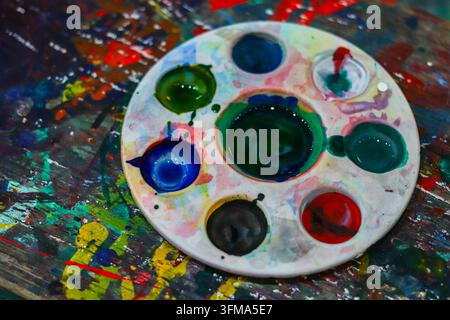 Weiße runde Farbpalette mit nasser Acrylfarbe auf einem bespritzten Holztisch in einer Kunstwerkstatt, hohe Winkelaufnahme, keine Personen. Stockfoto