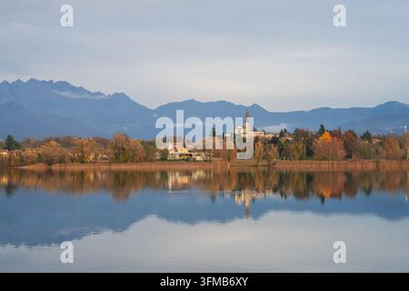 Der Pusiano-See, gelegen zwischen den Provinzen Como und Lecco in der Brianza, Italien, spiegelt Herbstbäume und ein entferntes Dorf wider, Bergkette unter sanftem Licht Stockfoto