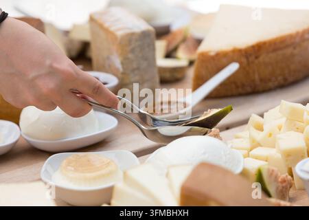 Hand, die mit einer Zange eine Feige von einem rustikalen Holzbrett mit verschiedenen Käsesorten serviert Stockfoto