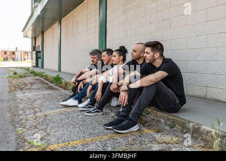 Gruppe verschiedener Freunde, die sich auf einer Bordsteinkante neben einem Gebäude in einer städtischen Gegend entspannen Stockfoto