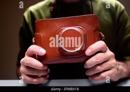neutrale Indooreinrichtung mit Händen, die eine Ledertasche für die Kamera im Nahaufnahme halten, Fotografie-Zubehör, braun und hellbraune Farben, natürliches Licht, sauber Stockfoto