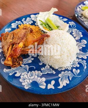 Köstliches indonesisches traditionelles Essen nasi bebek goreng, serviert mit gedämpftem weißem Reis, frischer Gurke, Kohl, Basilikumblättern und herzhaften gebratenen Flocken Stockfoto